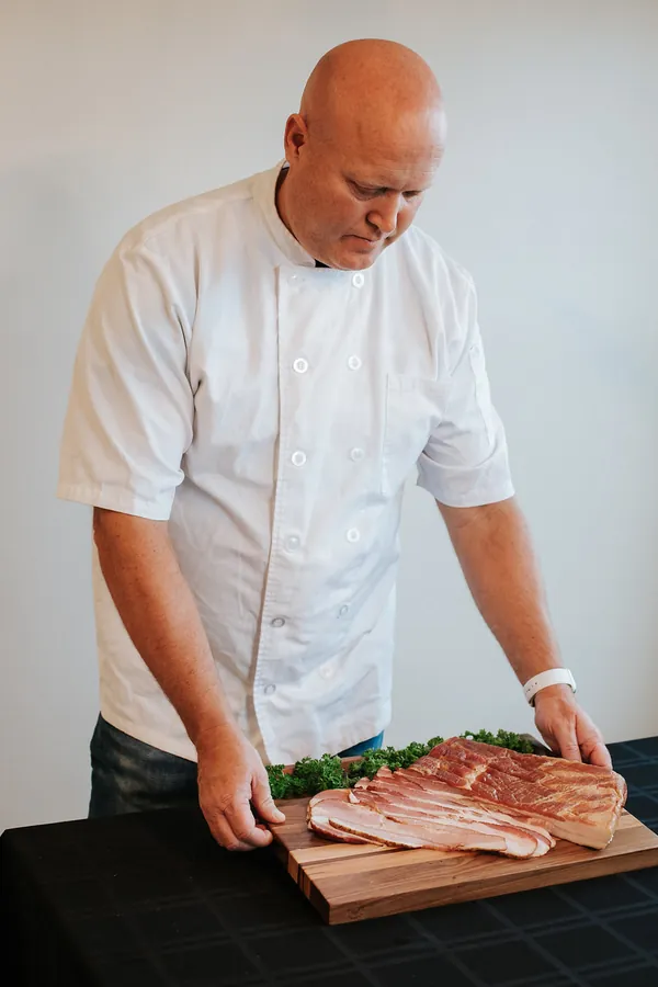 Tom Karren in a white chef coat setting down a board of sliced bacon